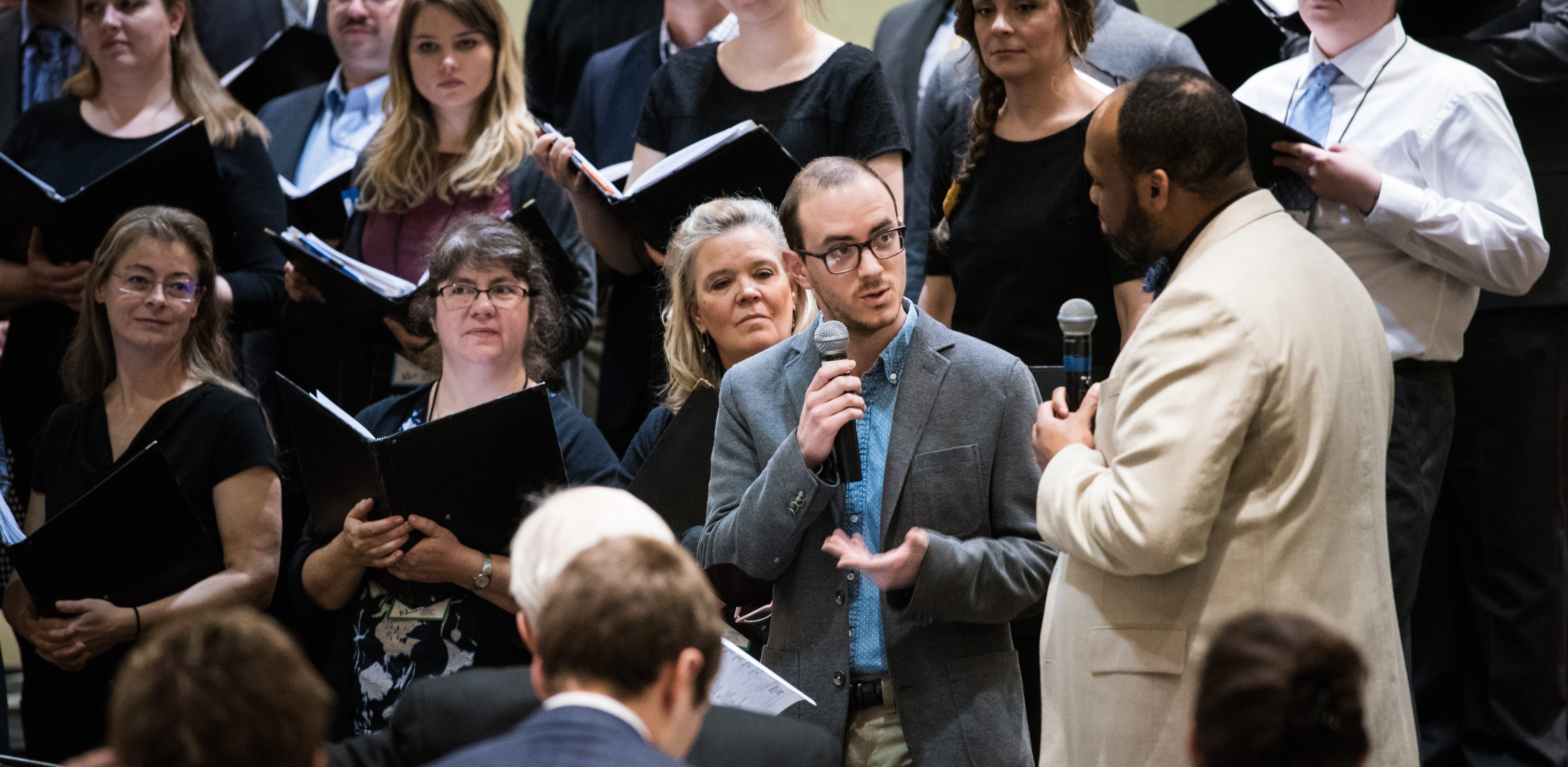 Two people with microphones talking in front of singers holding black folders. Photo credit: Bruce Silcox