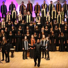 Conductor wearing black holds out hands in front of VocalEssence Singers wearing stoles. Photo Credit: Kyndell Harkness