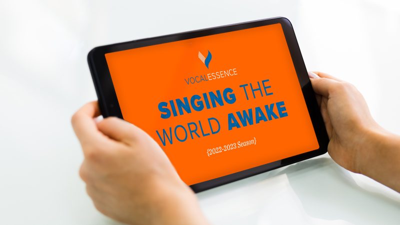 Hands holding tablet with VocalEssence logo, Singing the World Awake, and 2022-2023 Season on an orange background