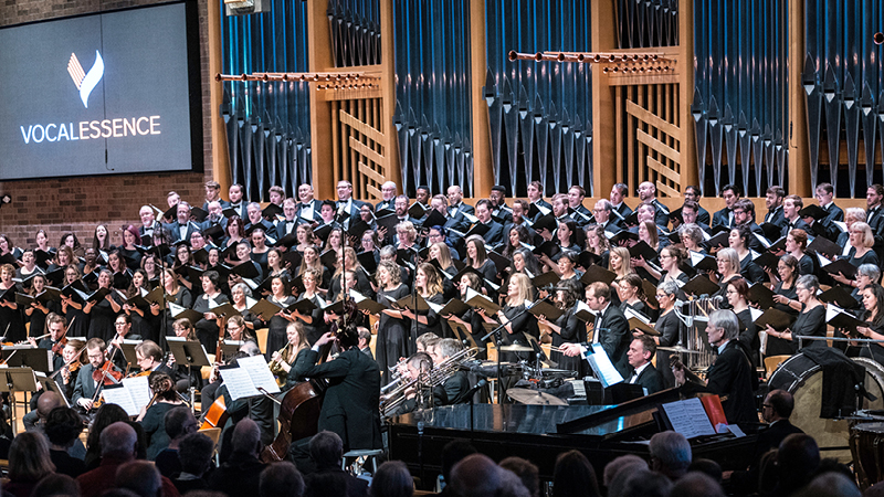 Members of VocalEssence holding black folders and singing with orchestra musicians in front of organ pipes with screen featuring VocalEssence logo in the upper left corner