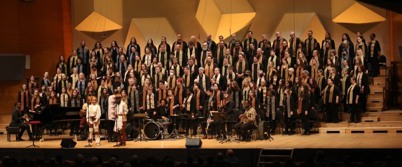 A large group of singers stands on the Orchestra Hall stage.