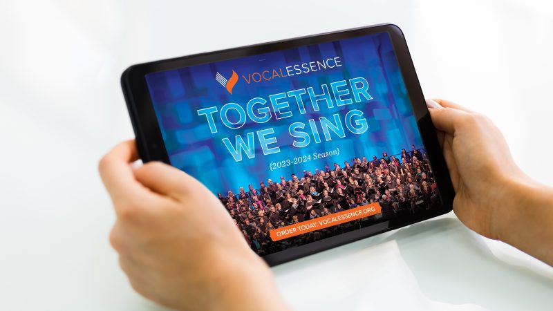 Hands holding tablet with VocalEssence logo, Together We Sing, 2023-2024 Season, with singers holding black folders performing at Northrop. Design: Lora Joshi, Photo Credit: Tony Nelson Photography