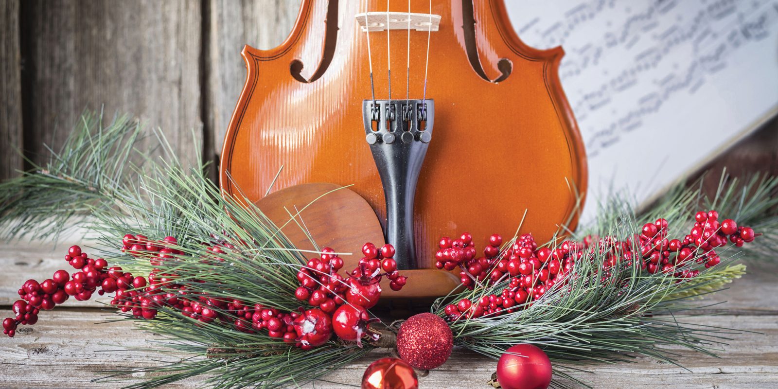 The bottom half of a violin with sheet music, pine, and cranberries adorning the front of the fiddle on rustic wood background. Photo Credit: Adobe Stock Image