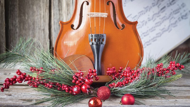The bottom half of a violin with sheet music, pine, and cranberries adorning the front of the fiddle on rustic wood background. Photo Credit: Adobe Stock Image