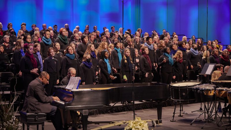 Person in black suit playing piano with singers wearing black and multicolored scarves singing
