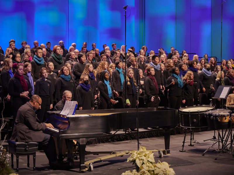 Person in black suit playing piano with singers wearing black and multicolored scarves singing