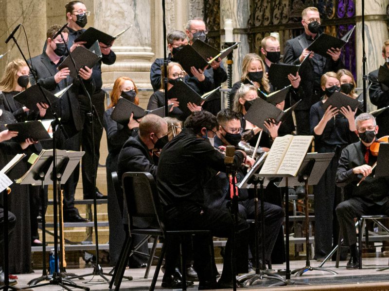 Conductor in black suit with red shirt in front of orchestra musicians and singers. Photo Credit: Bruce Silcox