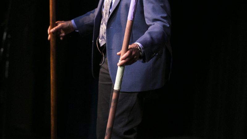 Melanie DeMore wears a blue suit and tie holding decorated pounding sticks. Photo Credit: Kyndell Harkness