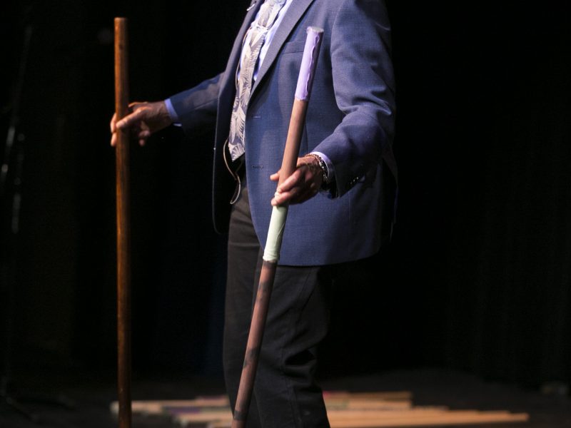Melanie DeMore wears a blue suit and tie holding decorated pounding sticks. Photo Credit: Kyndell Harkness