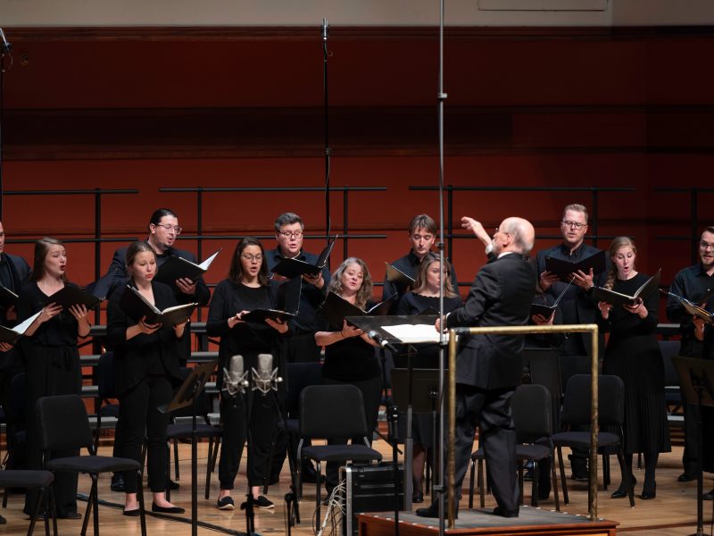 Conductor wearing black tuxedo in front of singers holding black folders and wearing black clothes. Photo Credit: Bruce Silcox