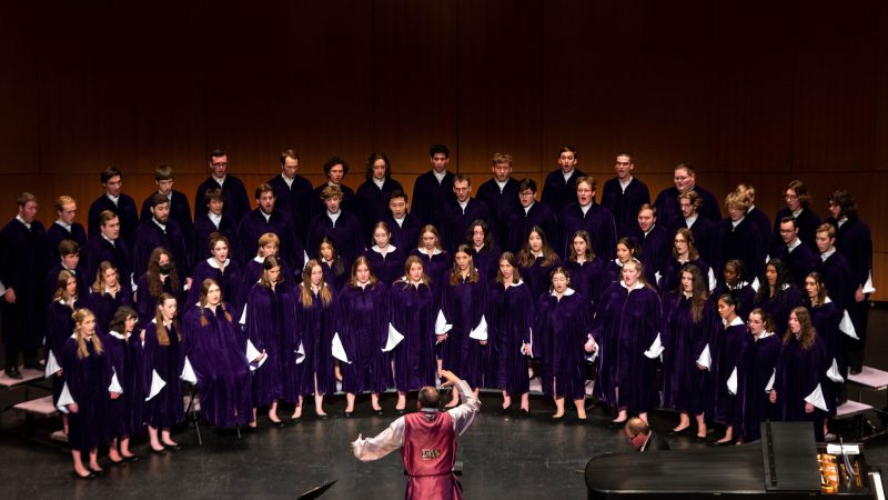 Conductor wearing a red robe in front of singers in blue robes