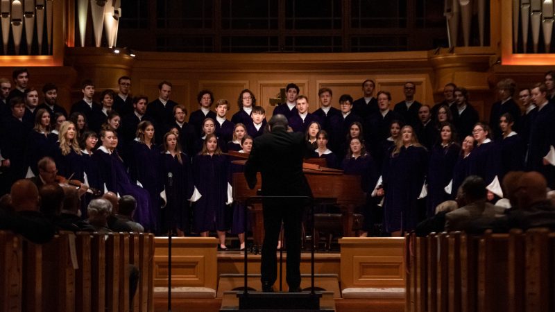 Conductor wearing black suit in front of singers in blue robes