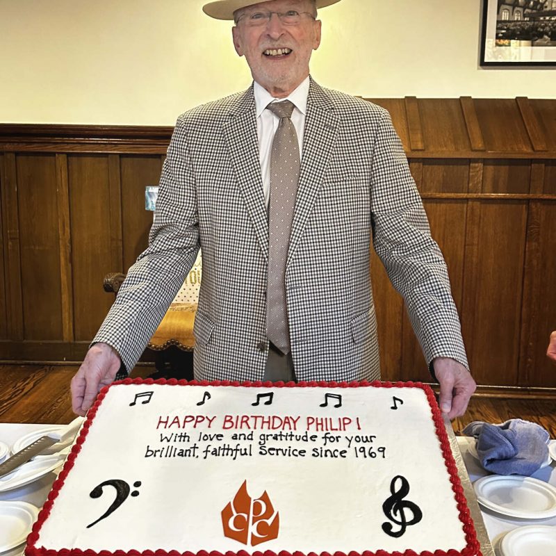 Philip Brunelle wearing a suit and a hat stands in front of a birthday cake.