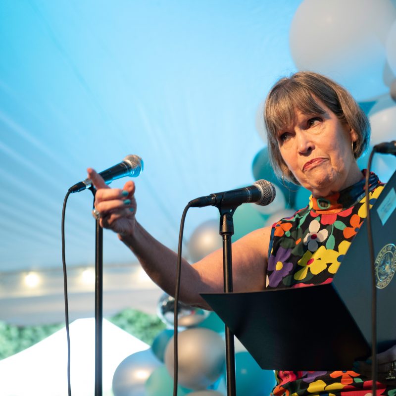 Representative Betty McCollum speaking into a microphone and gesturing with her pointer finger toward the audience in front of balloons.
