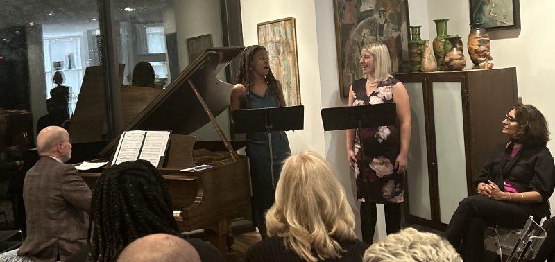 People sitting watching a person playing a piano and two people singing with black music stands in front of them. Photo credit: Elissa Weller.