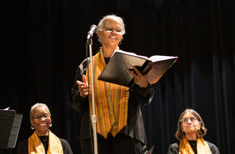 Person wearing glasses, a colorful stole, and holding a black folder speaks into a microphone with people smiling behind them. Photo credit-Nik Linde