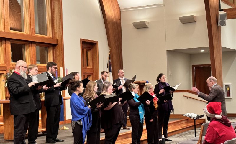 Singers with black folders performing with a conductor in front of them. Photo credit: Ethan Johnson