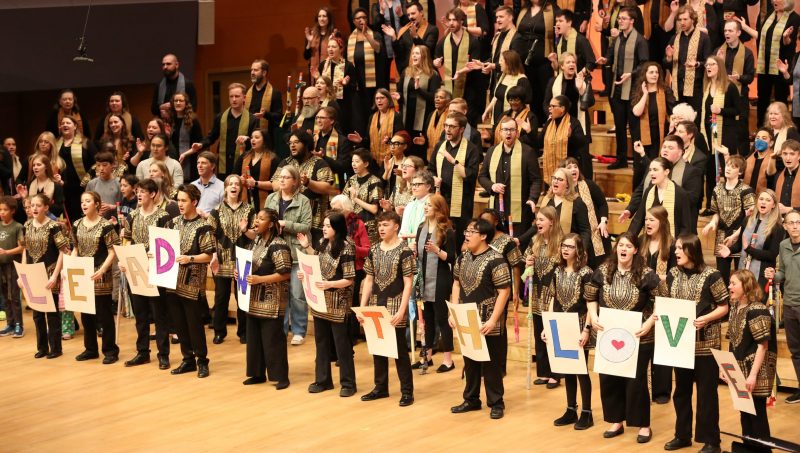 VocalEssence singers perform at Orchestra Hall. Several singers are holding signs with letters that read "LEAD WITH LOVE." Photo credit: Kyndell Harkness