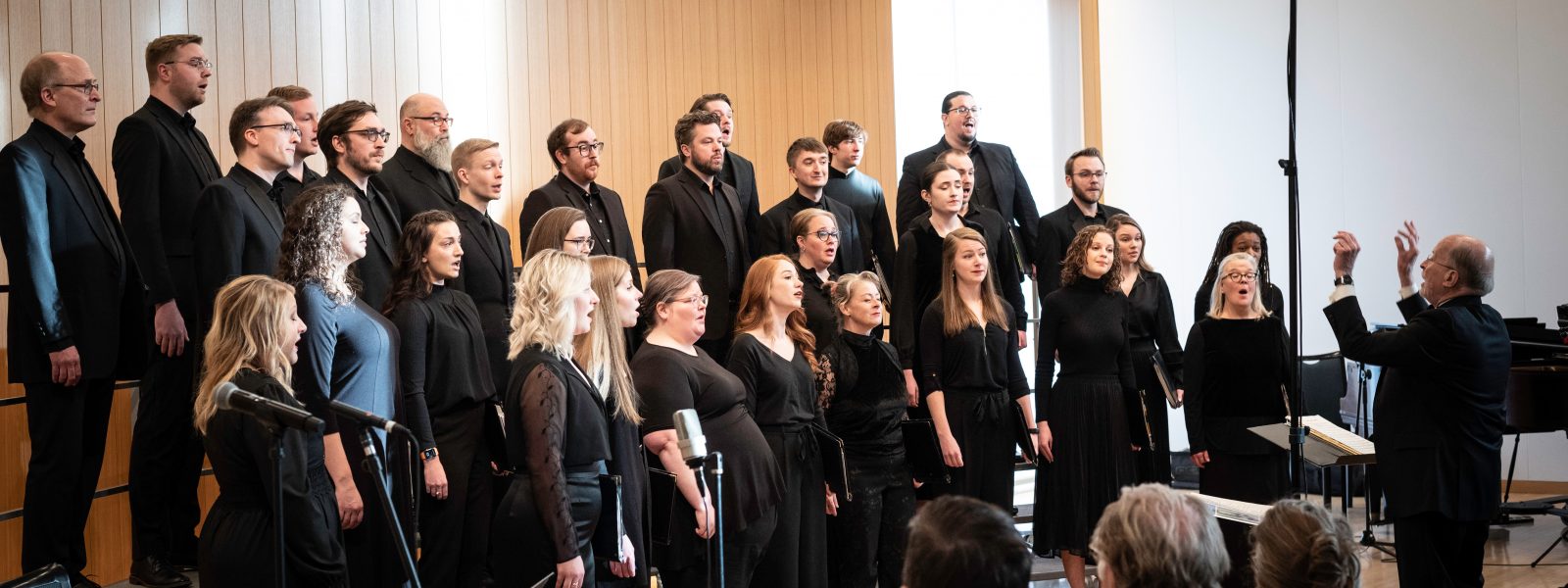 People dressed in black and holding black folders standing on risers in front of a paneled wall with a conductor leading them perform in front of an audience.