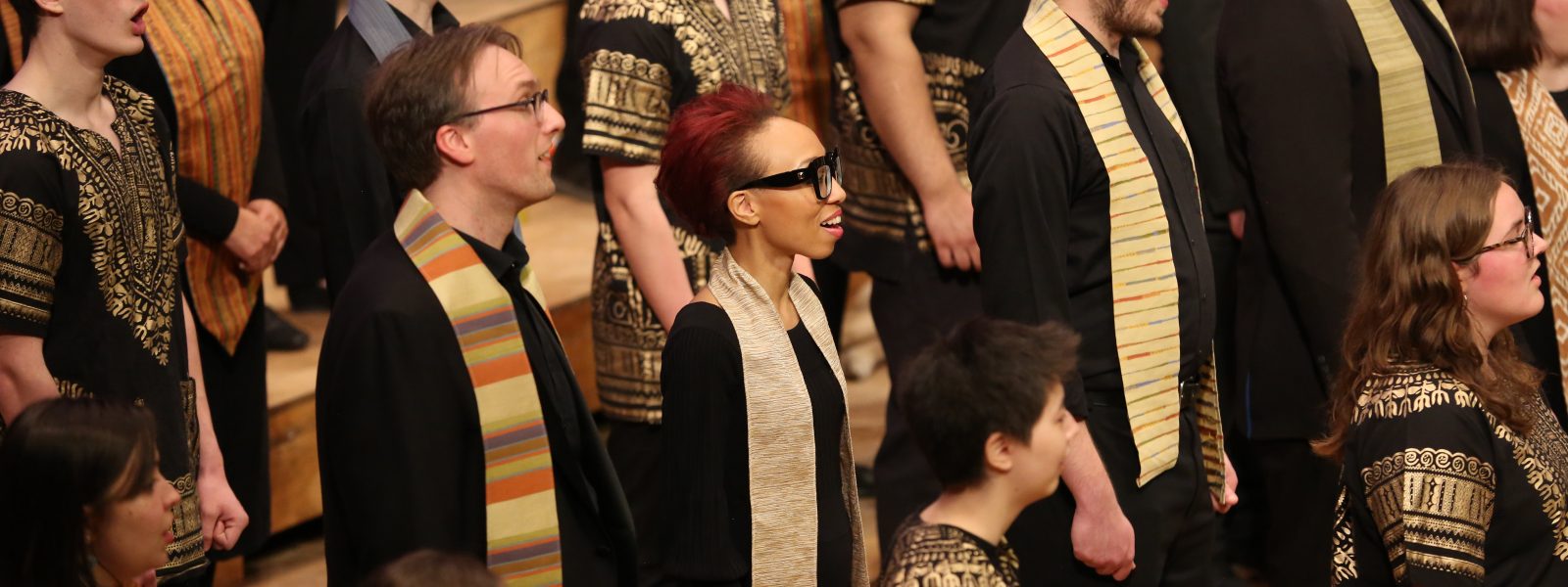 Singers wearing dark clothes with multicolored stoles sing.