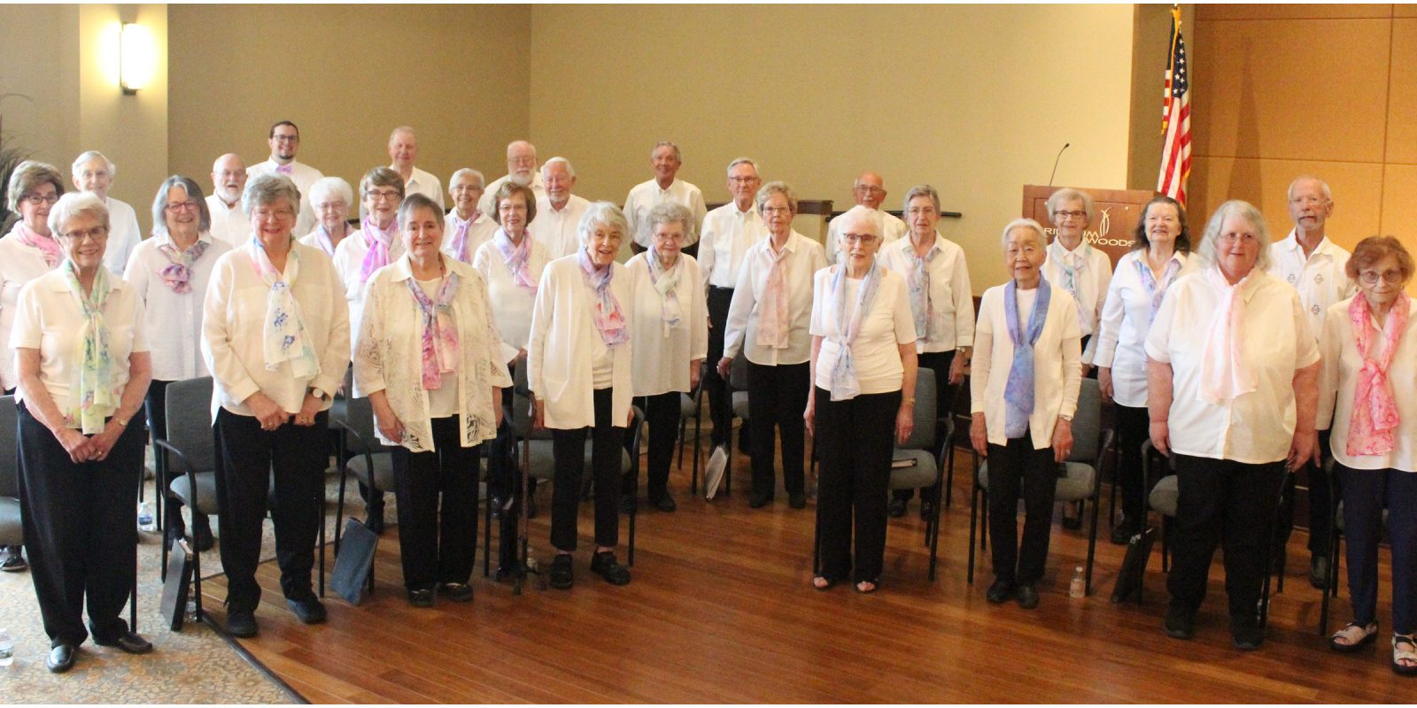 Trillium Woods Fall 2024 Event Page 2400x1200 Adult Vintage Voices members wearing white shirts, black pants standing, and spring colored scarves in a row and smiling. Photo Credit: Trillium Woods Staff