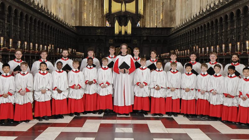 The Choir of King's College, Cambridge, comprised of boys and men, dressed in red cassocks with white surplices. They are standing in King's College Chapel, a masterpiece of Gothic architecture known for its soaring fan-vaulted ceiling, tall stained glass windows, and elaborate woodwork.