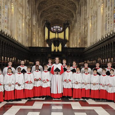 The Choir of King's College, Cambridge, comprised of boys and men, dressed in red cassocks with white surplices. They are standing in King's College Chapel, a masterpiece of Gothic architecture known for its soaring fan-vaulted ceiling, tall stained glass windows, and elaborate woodwork.