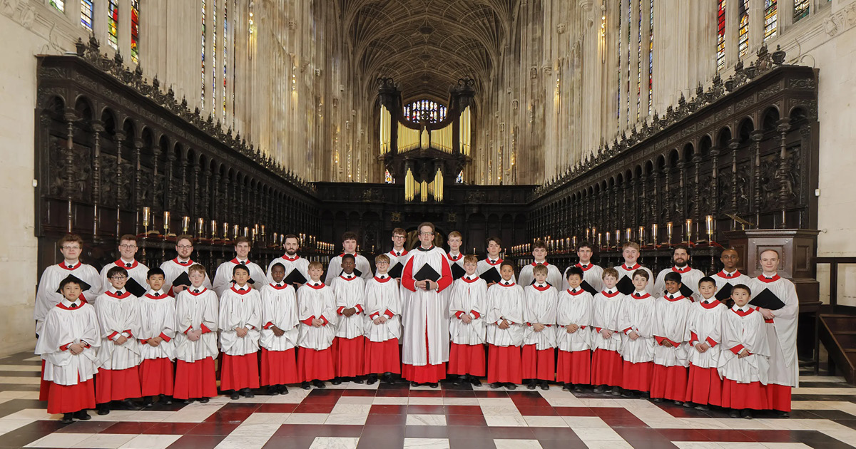 The Choir of King's College, Cambridge, comprised of boys and men, dressed in red cassocks with white surplices. They are standing in King's College Chapel, a masterpiece of Gothic architecture known for its soaring fan-vaulted ceiling, tall stained glass windows, and elaborate woodwork.