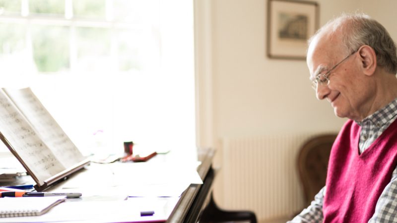John Rutter, wearing a red sweater vest over a checkered shirt, is smiling as he sits and plays the piano which has notebooks, pens, and sheet music. Photo credit: Unknown