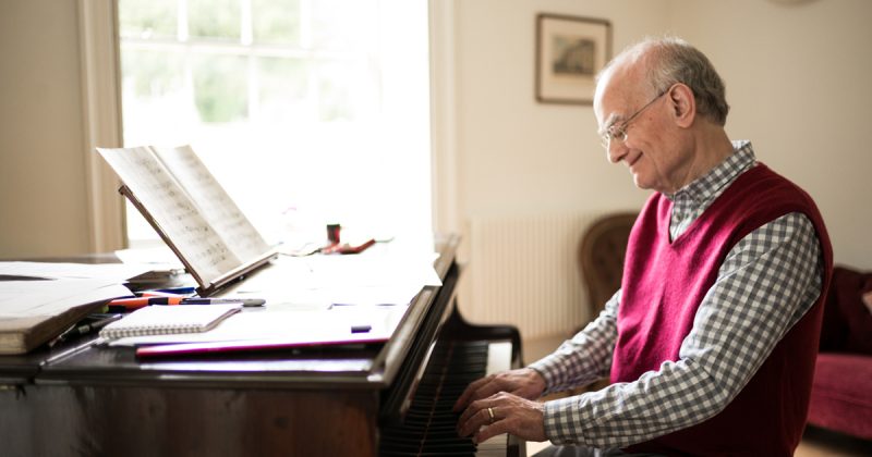 John Rutter, wearing a red sweater vest over a checkered shirt, is smiling as he sits and plays the piano which has notebooks, pens, and sheet music. Photo credit: Unknown