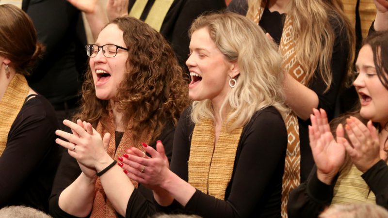 VocalEssence singers wearing black with multicolored stoles perform while clapping their hands with joyful expressions. Photo Credit: Kyndell Harkness