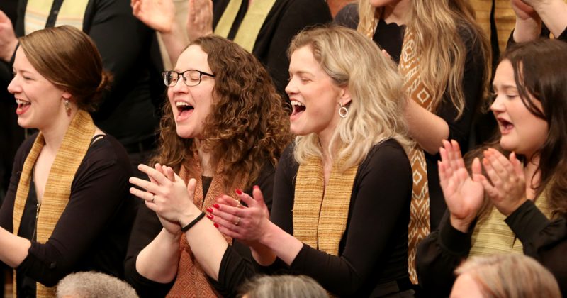 VocalEssence singers wearing black with multicolored stoles perform while clapping their hands with joyful expressions. Photo Credit: Kyndell Harkness