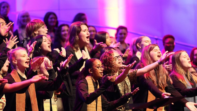 VocalEssence singers wearing black with multicolored stoles perform while holding their arms forward on stage for the annual WITNESS concert. Photo Credit: Kyndell Harkness
