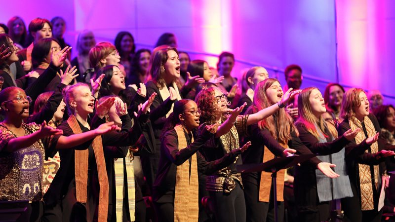 VocalEssence singers wearing black with multicolored stoles perform while holding their arms forward on stage for the annual WITNESS concert. Photo Credit: Kyndell Harkness