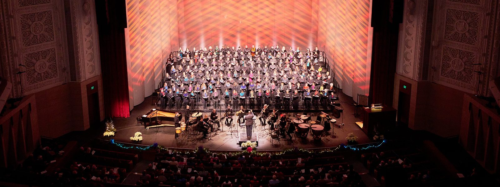 VocalEssence singers wearing black with colorful scarves holding black folders perform with brass and percussion on a poinsettia and twinkle light garland adorned stage at Northrop.