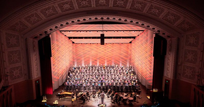 VocalEssence singers wearing black with colorful scarves holding black folders perform with brass and percussion on a poinsettia and twinkle light garland adorned stage at Northrop.