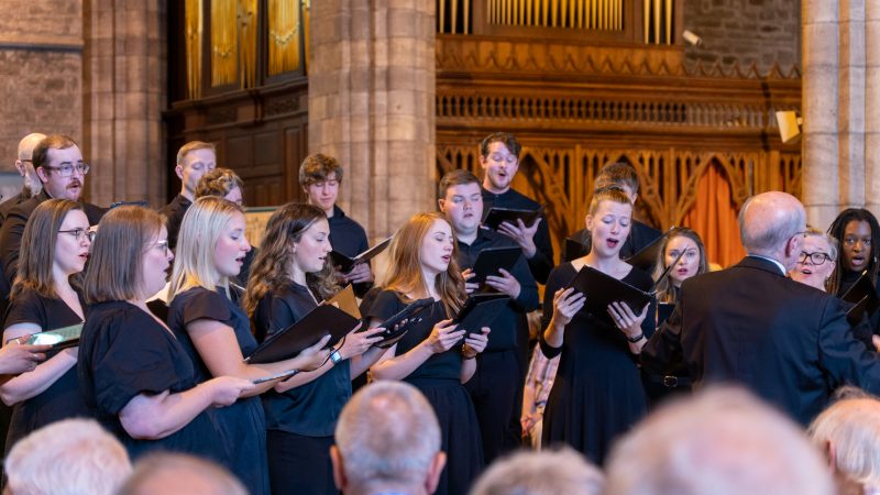 Philip Brunelle, wearing dark colored suits, conducts the VocalEssence Ensemble Singers wearing black holding folders and singing in front of an audience at a church at Three Choirs Festival. Photo Credit: Joseph Wong