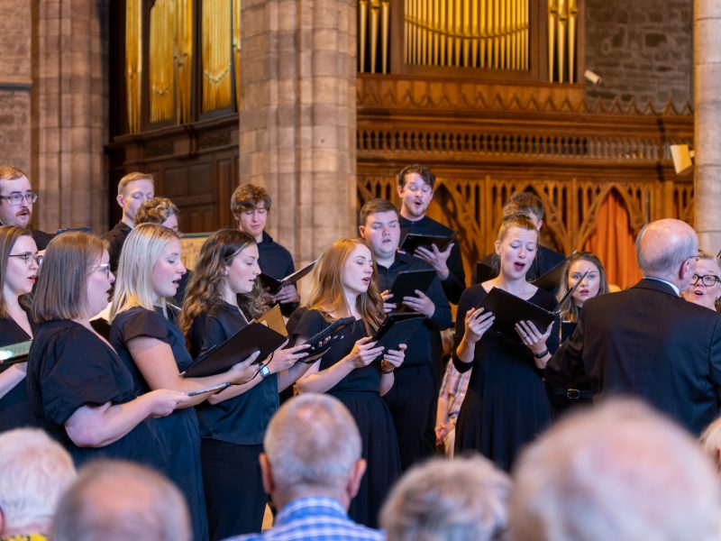 Philip Brunelle, wearing dark colored suits, conducts the VocalEssence Ensemble Singers wearing black holding folders and singing in front of an audience at a church at Three Choirs Festival. Photo Credit: Joseph Wong