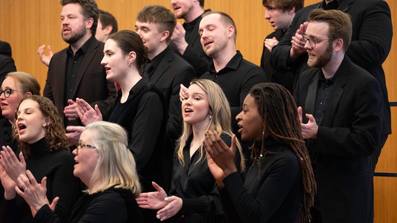 VocalEssence Ensemble Singers wearing black perform while clapping their hands with joyful expressions. Photo Credit: Bruce Silcox