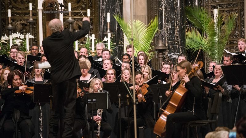 Person wearing a black suit conducts singers and orchestra wearing black. Photo credit: Bruce Silcox