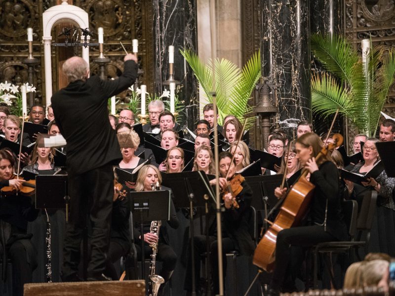 Person wearing a black suit conducts singers and orchestra wearing black. Photo credit: Bruce Silcox