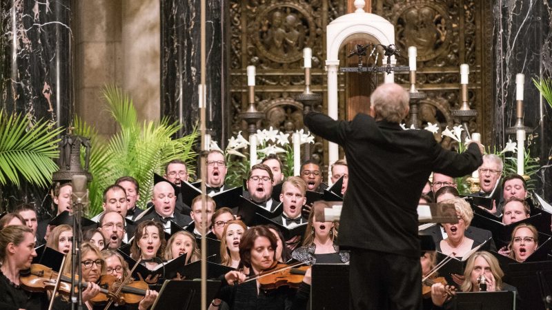 Person wearing a black suit conducts singers and orchestra wearing black. Photo credit: Bruce Silcox