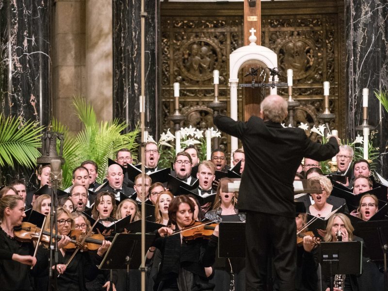 Person wearing a black suit conducts singers and orchestra wearing black. Photo credit: Bruce Silcox
