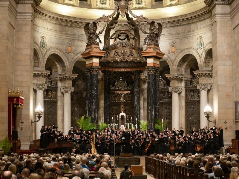 Many singers perform in front of an audience in the ornate sanctuary of the Cathedral of Saint Paul that features a baldachin with black and gold marble columns that has a bronze latticework canopy. Photo Credit: Bruce Silcox