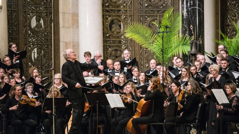 Person wearing a black suit conducts singers and orchestra wearing black. Photo credit: Bruce Silcox