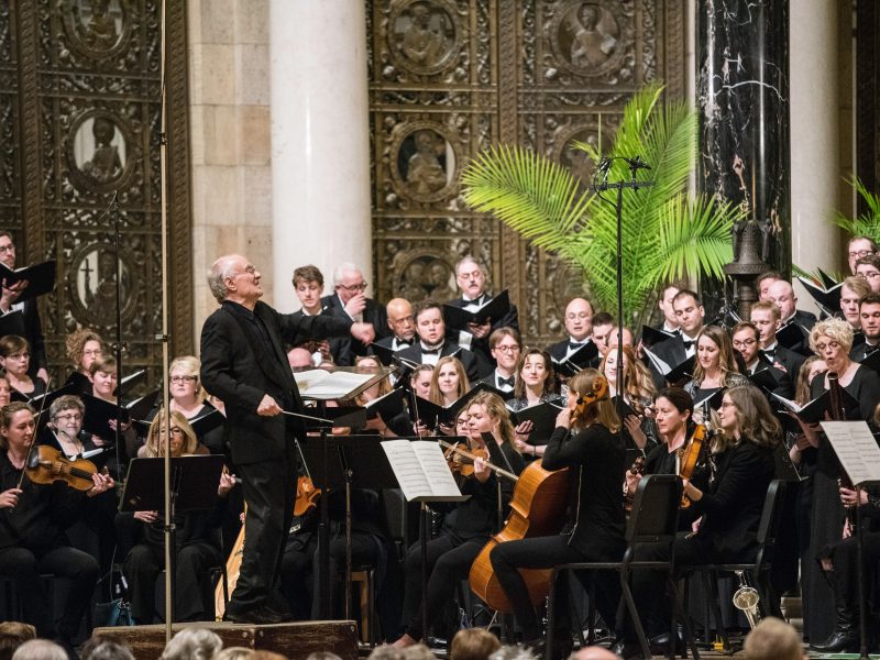 Person wearing a black suit conducts singers and orchestra wearing black. Photo credit: Bruce Silcox