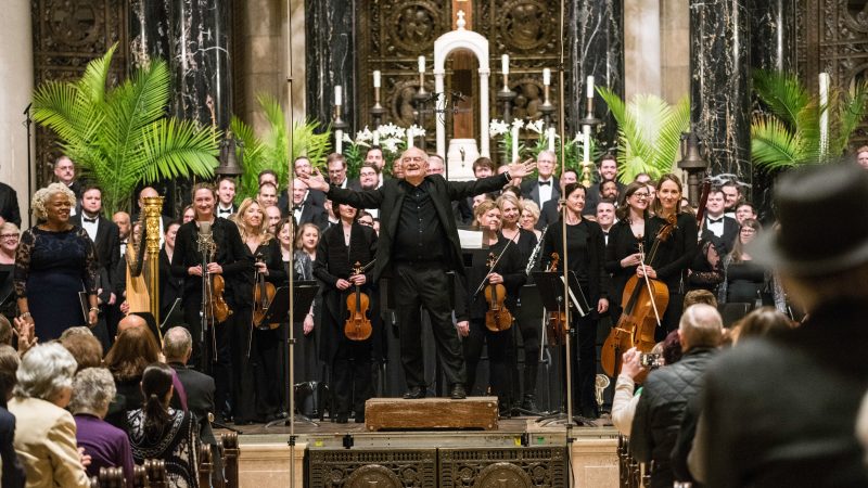 Person wearing black suit with arms outstretched stands in front of singers and orchestra in front of an audience in the ornate sanctuary of the Cathedral of Saint Paul. Photo Credit: Bruce Silcox