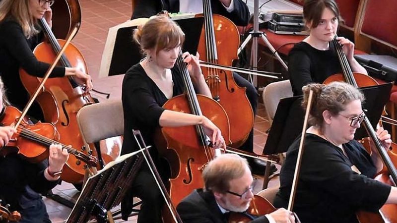 Members of the Metropolitan Symphony Orchestra wearing black, sitting, playing their instruments, with black music stands.