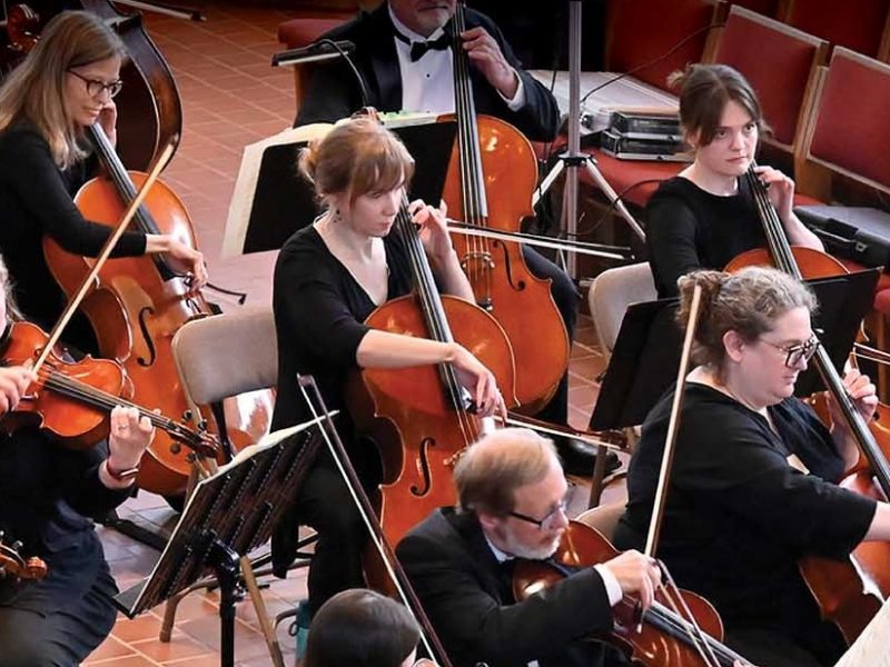Members of the Metropolitan Symphony Orchestra wearing black, sitting, playing their instruments, with black music stands.