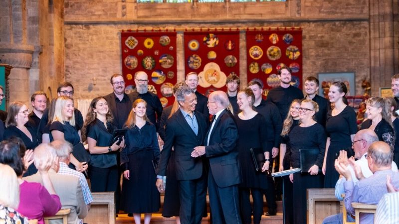 Roderick Williams and Philip Brunelle smiling and wearing dark colored suits shake hands in front of VocalEssence Ensemble Singers wearing black and standing in front of an audience at a church at Three Choirs Festival. Photo Credit: Joseph Wong